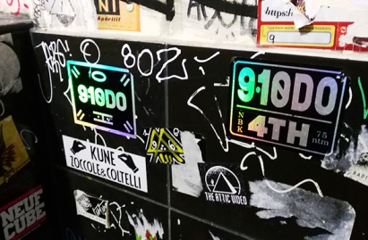 Collage de stickers holographiques aux couleurs vives et designs variés imprimés, apposés sur une surface urbaine évoquant une impression typique de l'art de rue.